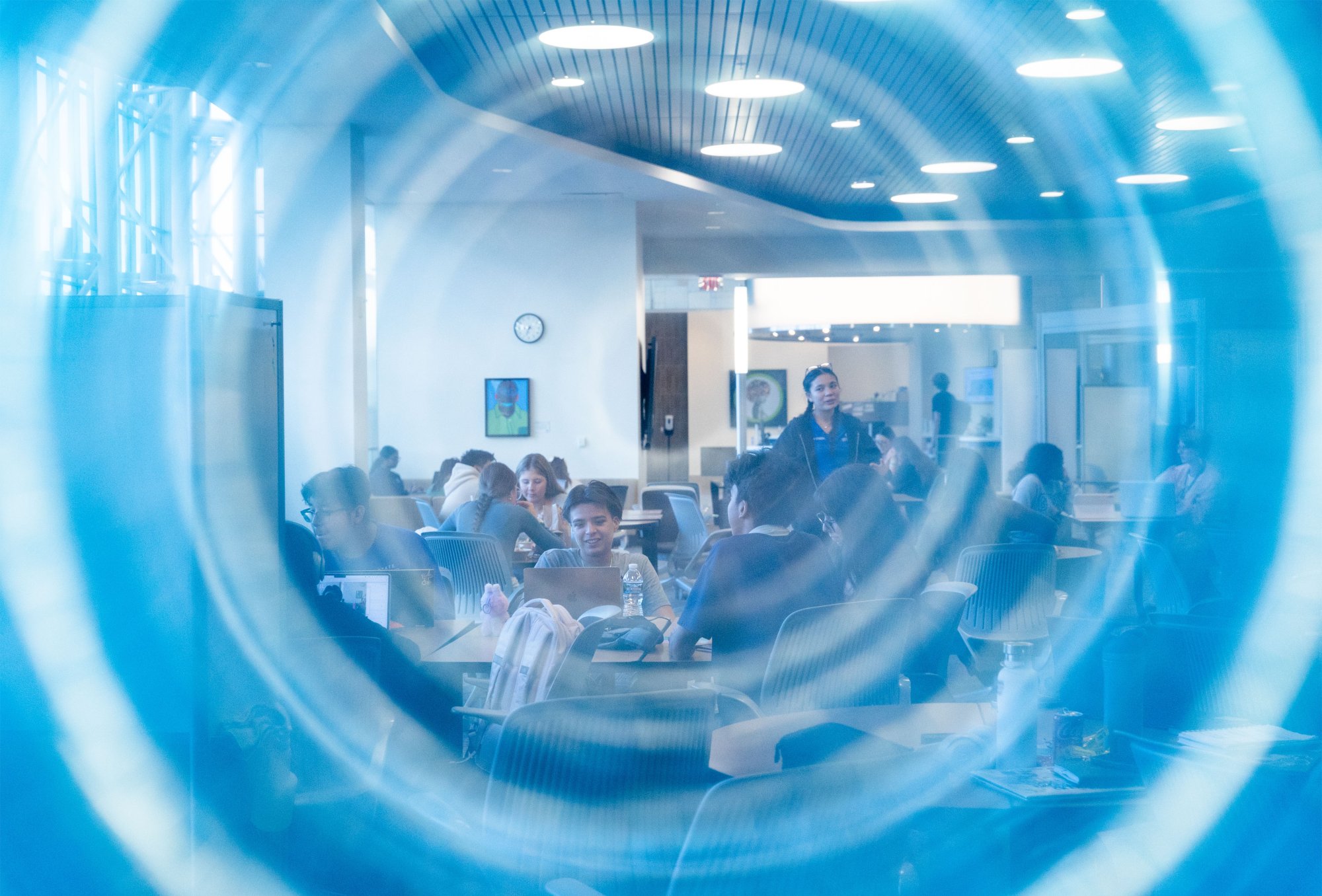 A group of students gather in a bright, modern study area. Blue circular shapes overlay the scene, adding an artistic, dynamic feel. The atmosphere is focused and collaborative